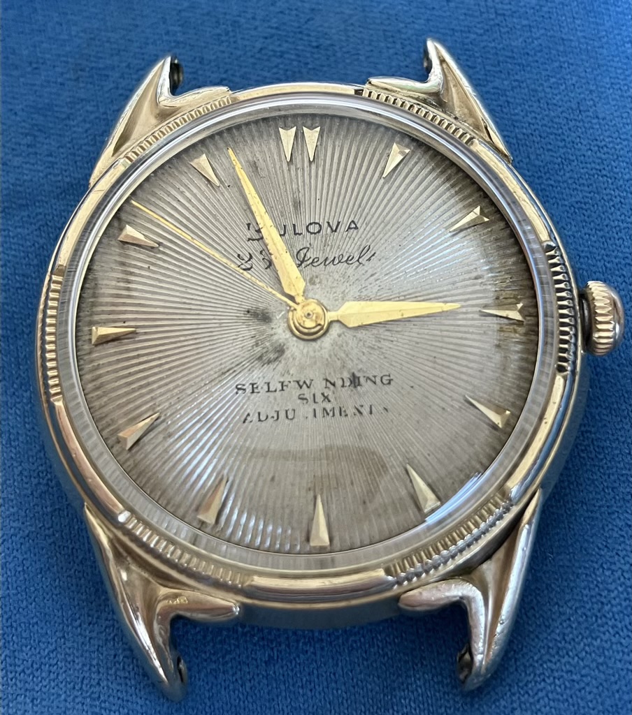 Vintage Bulova self-winding wristwatch with gold-tone hands and triangular hour markers on a sunburst dial. The case is gold-toned and shows wear, laid on a blue fabric background.