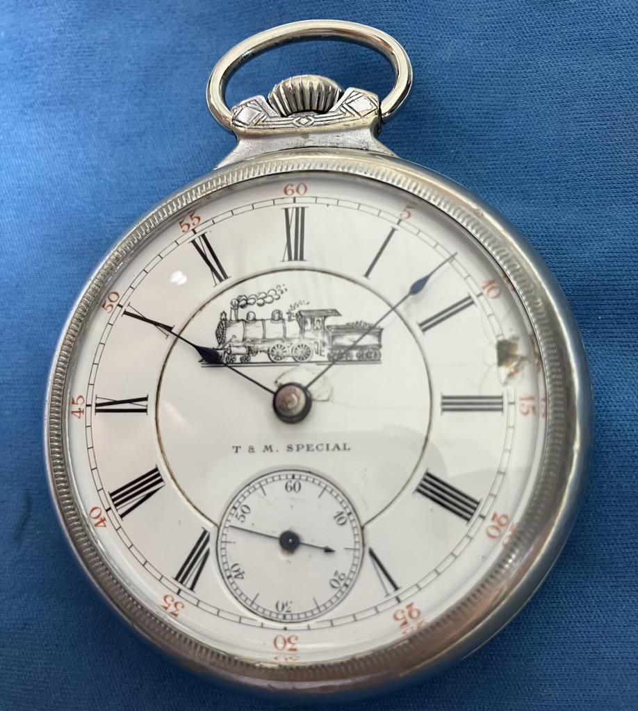 Silver pocket watch with Roman numerals and a train engraving, labeled 'T & M Special' on a blue background