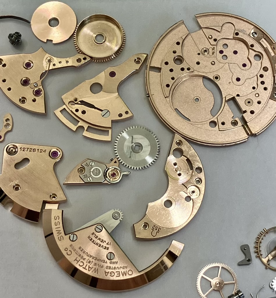 Assorted brass watch movement parts and gears laid out on a gray surface, including plates and jeweled bearings.