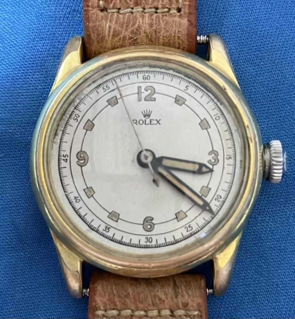 Vintage Rolex wristwatch with a brown leather strap on a blue background; round gold case and white dial with Arabic numerals 12,3,6,9 and inner minute ring